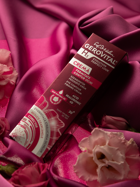 Gerovital Eye Contour Firming Cream box on burgundy fabric beside pink flowers